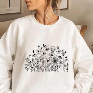 Op de afbeelding: Witte trui met een zwarte lijntekening van een bloemenveld met vlinders.