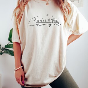 May include: A white oversized t-shirt with a black graphic design that says "Happy Camper" with a camper van, trees, and stars.