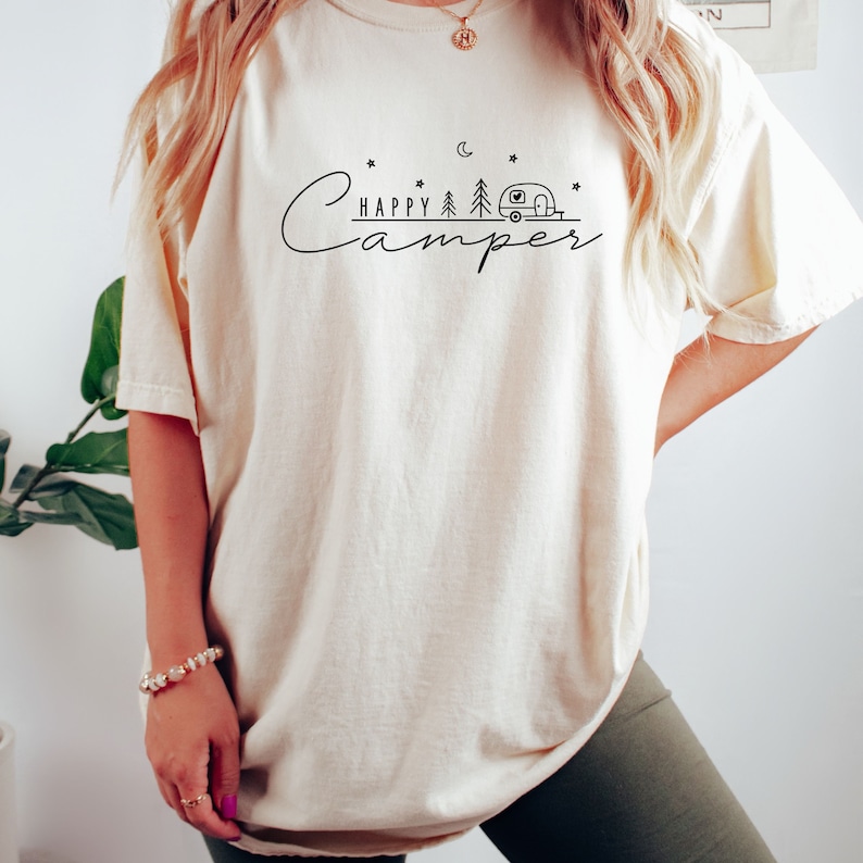 May include: A white oversized t-shirt with a black graphic design that says "Happy Camper" with a camper van, trees, and stars.