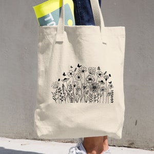 Op de afbeelding: Een witte canvas tote bag met zwarte lijnkunst van een veld met bloemen en vlinders. De tas heeft twee lange handvatten.