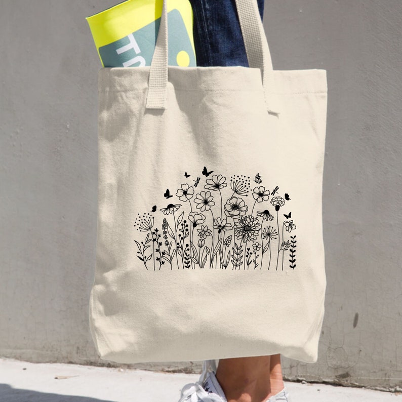 Op de afbeelding: Een witte canvas tote bag met zwarte lijnkunst van een veld met bloemen en vlinders. De tas heeft twee lange handvatten.