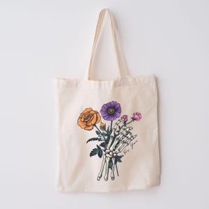 May include: A white tote bag with a skeleton hand holding a bouquet of flowers. The flowers are orange, purple, and pink. The text "For You" is written on the bag.