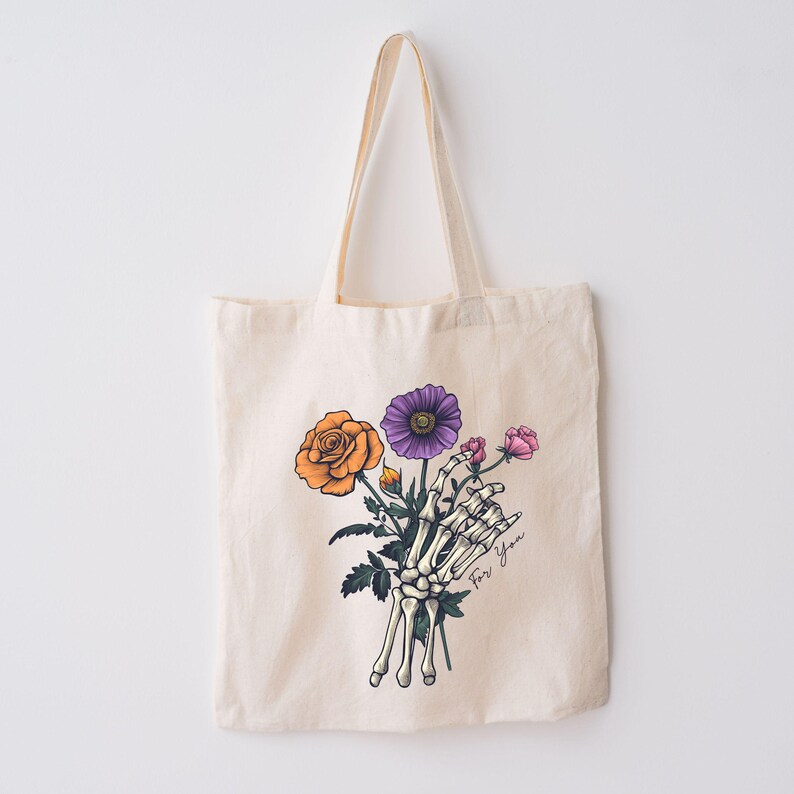 May include: A white tote bag with a skeleton hand holding a bouquet of flowers. The flowers are orange, purple, and pink. The text "For You" is written on the bag.