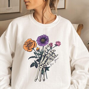 May include: White sweatshirt with a graphic of a skeleton hand holding a bouquet of flowers. The flowers are orange, purple, and pink. The text "For You" is written on the sweatshirt.