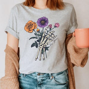 May include: A gray t-shirt with a skeleton hand holding a bouquet of flowers. The flowers are orange, purple, and pink. The text "For You" is written on the shirt.