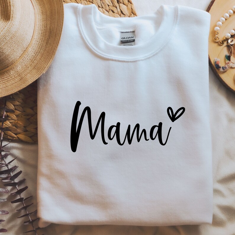 Może przedstawiać: Biała bluza z napisem "Mama" napisanym czarnym, ozdobnym pismem z małym sercem obok.