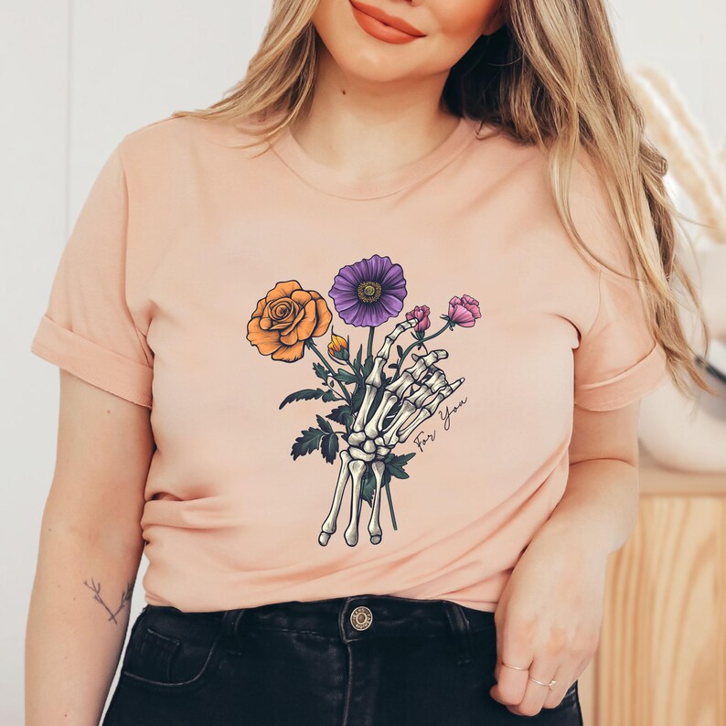 May include: A peach-colored t-shirt with a skeleton hand holding a bouquet of flowers. The flowers are orange, purple, and pink. The text "For You" is written on the shirt.