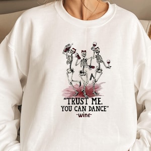 Peut inclure: Sweat-shirt blanc avec un motif de trois squelettes dansant, tenant des bouteilles et des verres de vin. Le texte "TRUST ME. YOU CAN DANCE" et "-wine-" est imprim&eacute; en dessous.