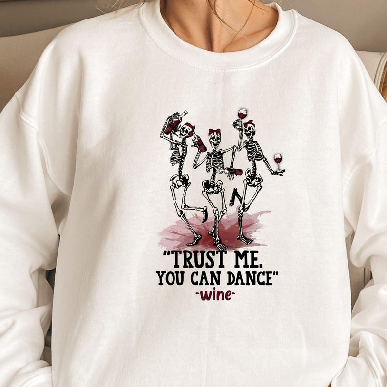 Peut inclure: Sweat-shirt blanc avec un motif de trois squelettes dansant, tenant des bouteilles et des verres de vin. Le texte "TRUST ME. YOU CAN DANCE" et "-wine-" est imprim&eacute; en dessous.