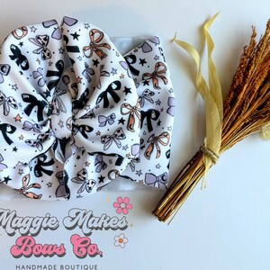 May include: A white baby headband with a pattern of black, lavender, and peach bows and stars. The headband is knotted in the center. The image also includes a bundle of dried flowers and the text "Maggie Makes Bows Co. HANDMADE BOUTIQUE."