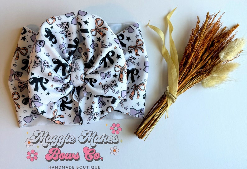 May include: A white baby headband with a pattern of black, lavender, and peach bows and stars. The headband is knotted in the center. The image also includes a bundle of dried flowers and the text "Maggie Makes Bows Co. HANDMADE BOUTIQUE."