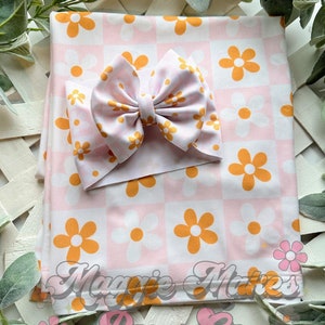 May include: A pink and orange floral print baby blanket with a matching pink and white bow headband. The blanket has a checkered pattern with orange and white flowers. The headband is tied in a bow and has a white flower pattern.
