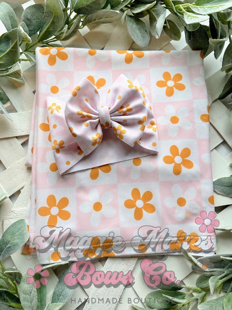May include: A pink and orange floral print baby blanket with a matching pink and white bow headband. The blanket has a checkered pattern with orange and white flowers. The headband is tied in a bow and has a white flower pattern.