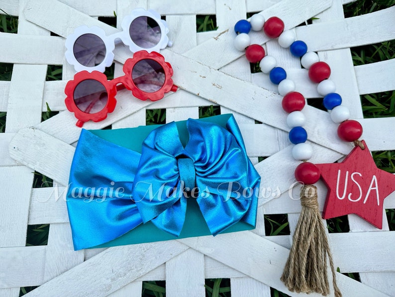 May include: A turquoise blue headband with a large bow, red and white flower-shaped sunglasses, and a red, white, and blue beaded garland with a wooden star that says "USA".