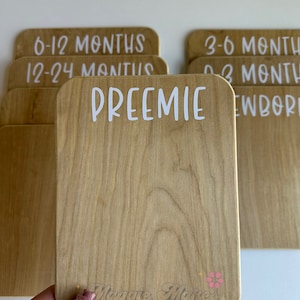 May include: Wooden milestone cards with rounded corners and white text. One card reads "PREEMIE", others read "0-3 MONTHS", "3-6 MONTHS", "6-12 MONTHS", "12-24 MONTHS", and "NEWBORN". The cards are for baby photos.
