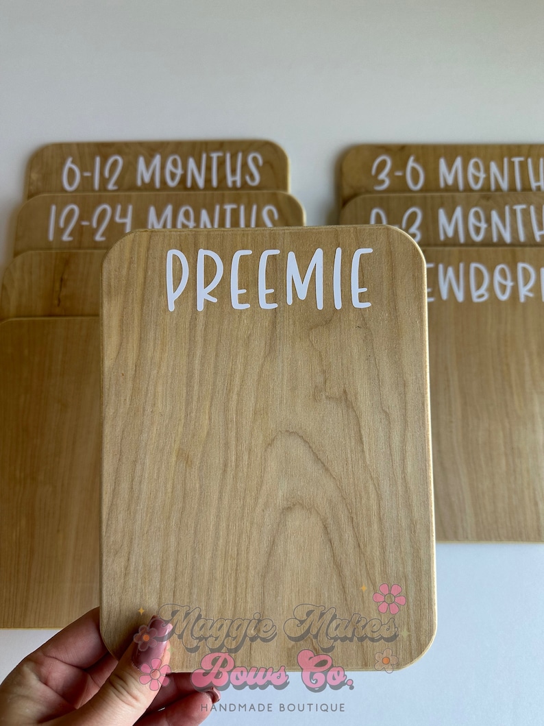 May include: Wooden milestone cards with rounded corners and white text. One card reads "PREEMIE", others read "0-3 MONTHS", "3-6 MONTHS", "6-12 MONTHS", "12-24 MONTHS", and "NEWBORN". The cards are for baby photos.