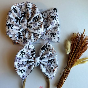 May include: A set of three baby hair accessories: a large bow on a headband, a matching bow on a nylon band, and a wide headband. All are white with a repeating pattern of black, orange, and lavender bows and stars. The brand name "Maggie Makes Bows Co." is printed at the bottom.