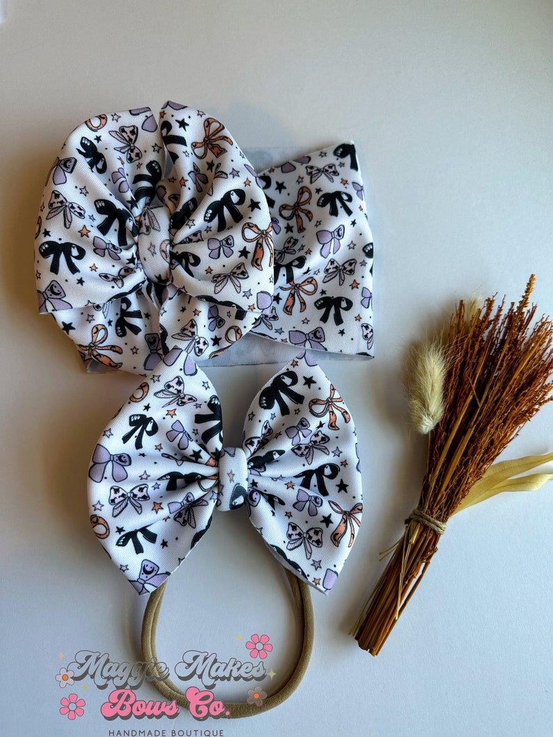 May include: A set of three baby hair accessories: a large bow on a headband, a matching bow on a nylon band, and a wide headband. All are white with a repeating pattern of black, orange, and lavender bows and stars. The brand name "Maggie Makes Bows Co." is printed at the bottom.