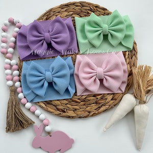 May include: Four pastel-colored fabric headbands with large bows. The headbands are arranged in a circle on a woven straw mat. The colors are lavender, light green, light blue, and pink.
