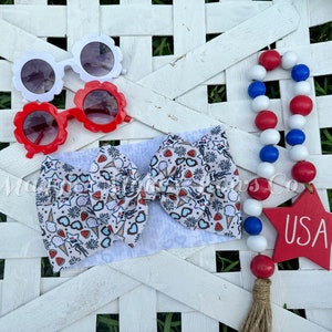 May include: A white headband with a red, white, and blue ice cream and fireworks print. The headband is tied in a bow. There are also red and white sunglasses, a red star with the word "USA" on it, and a red, white, and blue beaded garland.