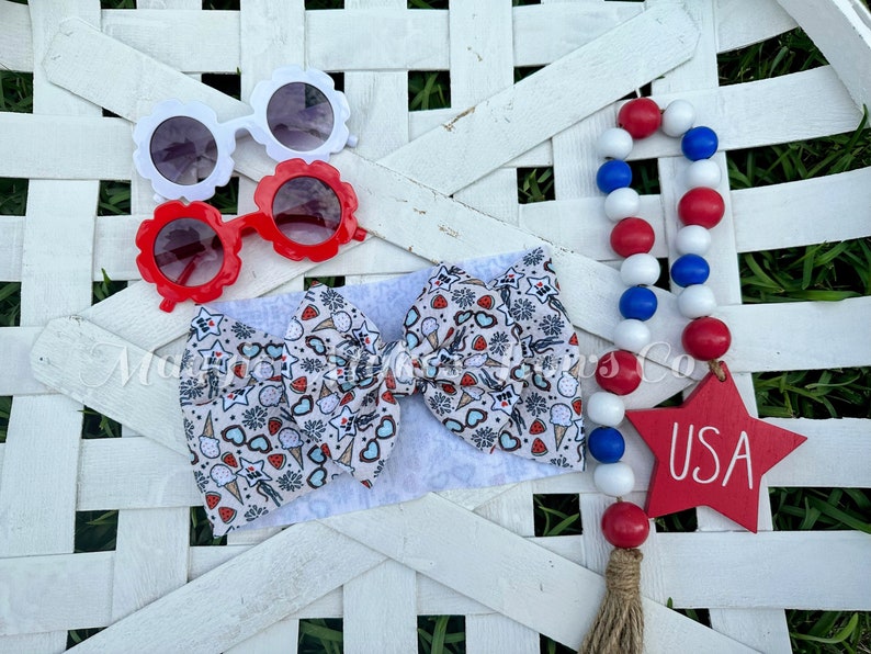 May include: A white headband with a red, white, and blue ice cream and fireworks print. The headband is tied in a bow. There are also red and white sunglasses, a red star with the word "USA" on it, and a red, white, and blue beaded garland.