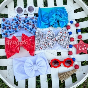 May include: A collection of red, white, and blue hair bows and sunglasses, perfect for a patriotic celebration. The bows are made of different fabrics, including satin, cotton, and patterned fabric. The sunglasses are red and white with a flower-shaped frame. The image also includes a red, white, and blue beaded garland with a star-shaped wooden sign that says "USA".
