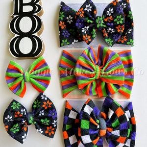 May include: Four Halloween-themed hair bows with different patterns: black with white and green flowers, orange and green stripes, black and white checkered, and orange, green, black, and white checkered. The word "BOO" is written in black on a white wooden sign.