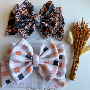 May include: Two fabric bows. The top bow is black with a floral and "Hocus Pocus" print in orange and pink. The bottom bow is white with a plaid pattern in orange, pink, and black. The bows are on white headbands.