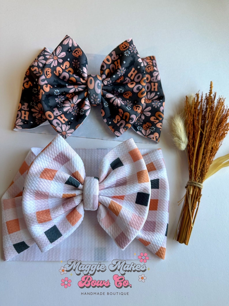 May include: Two fabric bows. The top bow is black with a floral and "Hocus Pocus" print in orange and pink. The bottom bow is white with a plaid pattern in orange, pink, and black. The bows are on white headbands.