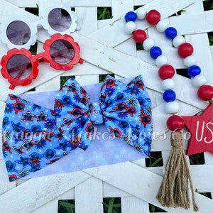 May include: A blue and white headband with a bow featuring a red, white, and blue sunglasses pattern. The headband is accompanied by a red, white, and blue beaded garland with a red wooden star that says "USA" and a brown tassel.