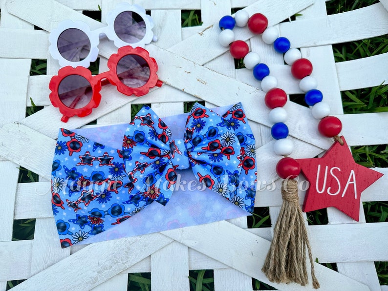 May include: A blue and white headband with a bow featuring a red, white, and blue sunglasses pattern. The headband is accompanied by a red, white, and blue beaded garland with a red wooden star that says "USA" and a brown tassel.