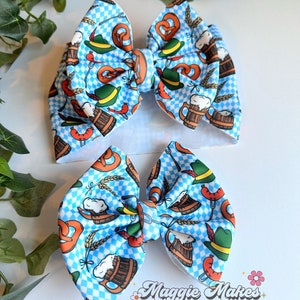 May include: Two fabric hair bows with a blue and white checkered pattern. The bows are decorated with images of pretzels, beer steins, sausages, and green hats. The bows are part of a set and are designed for babies or toddlers.