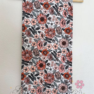 May include: A white fabric with a floral print featuring pink, orange, and black flowers. The fabric is hanging on a wooden hanger.