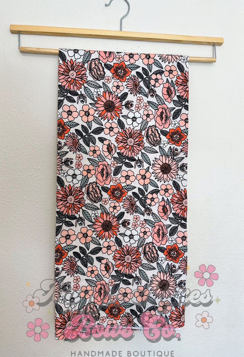 May include: A white fabric with a floral print featuring pink, orange, and black flowers. The fabric is hanging on a wooden hanger.