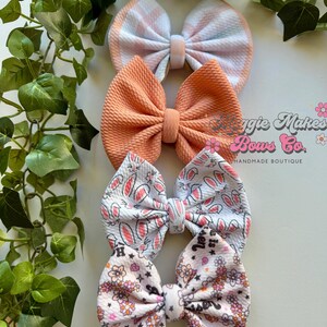 May include: Four handmade hair bows in various patterns. The top bow is white and light blue with a checkered pattern. The second bow is a solid orange color. The third bow is white with a pink bunny rabbit pattern. The bottom bow is white with a pink floral pattern and the text "Love is Sweet".