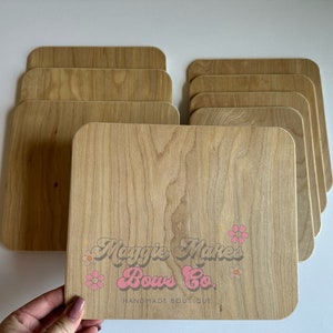 May include: A stack of light-colored wooden cutting boards with rounded corners. The boards have a natural wood grain pattern. The bottom board has the text "Maggie's Makes Bows Co. Handmade Boutique" in pink and orange.