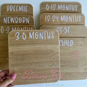 May include: A collection of wooden milestone plaques with rounded corners. Each plaque features white text indicating age ranges such as "Preemie," "Newborn," "0-3 Months," "3-6 Months," "6-12 Months," "12-24 Months," "Child." The bottom plaque has the text "Maggie's Makes Bows Co."