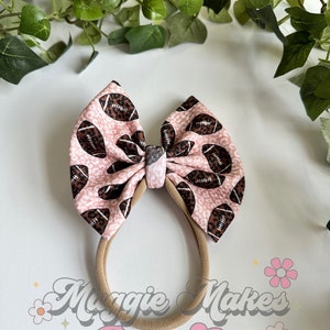 May include: A pink and brown headband with a bow featuring a football pattern. The headband is made of elastic and has a brown elastic band. The bow is tied with a brown ribbon. The text "Maggie Makes Bows Co. HANDMADE BOUTIQUE" is visible in the image.