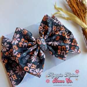 May include: A large, handmade hair bow featuring a dark gray background with a floral and Halloween-themed pattern. The bow has the words "Hocus Pocus" in orange and pink, along with pumpkins and flowers. The bow is from Maggie Makes Bows Co.