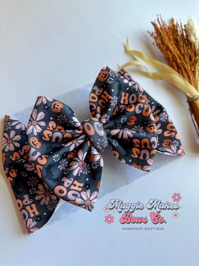 May include: A large, handmade hair bow featuring a dark gray background with a floral and Halloween-themed pattern. The bow has the words "Hocus Pocus" in orange and pink, along with pumpkins and flowers. The bow is from Maggie Makes Bows Co.
