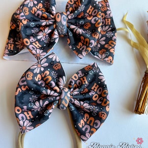 May include: Two Halloween-themed hair bows. The bows feature a black background with a floral pattern and the words "Hocus Pocus" in orange and pink. One bow is attached to a headband.