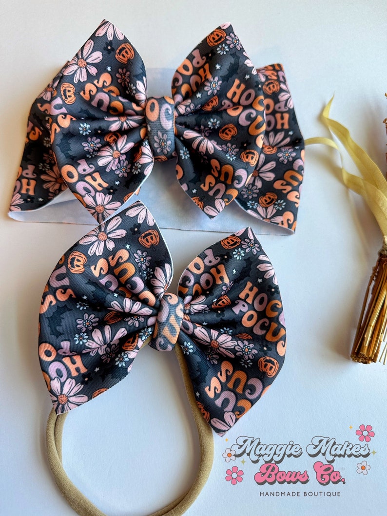 May include: Two Halloween-themed hair bows. The bows feature a black background with a floral pattern and the words "Hocus Pocus" in orange and pink. One bow is attached to a headband.