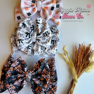 May include: Three fabric headbands with bow accents. The top headband has a pink, orange, and black checkered pattern. The middle headband is white with black and lavender bows. The bottom headband is black with orange flowers and the text "Hocus Pocus".