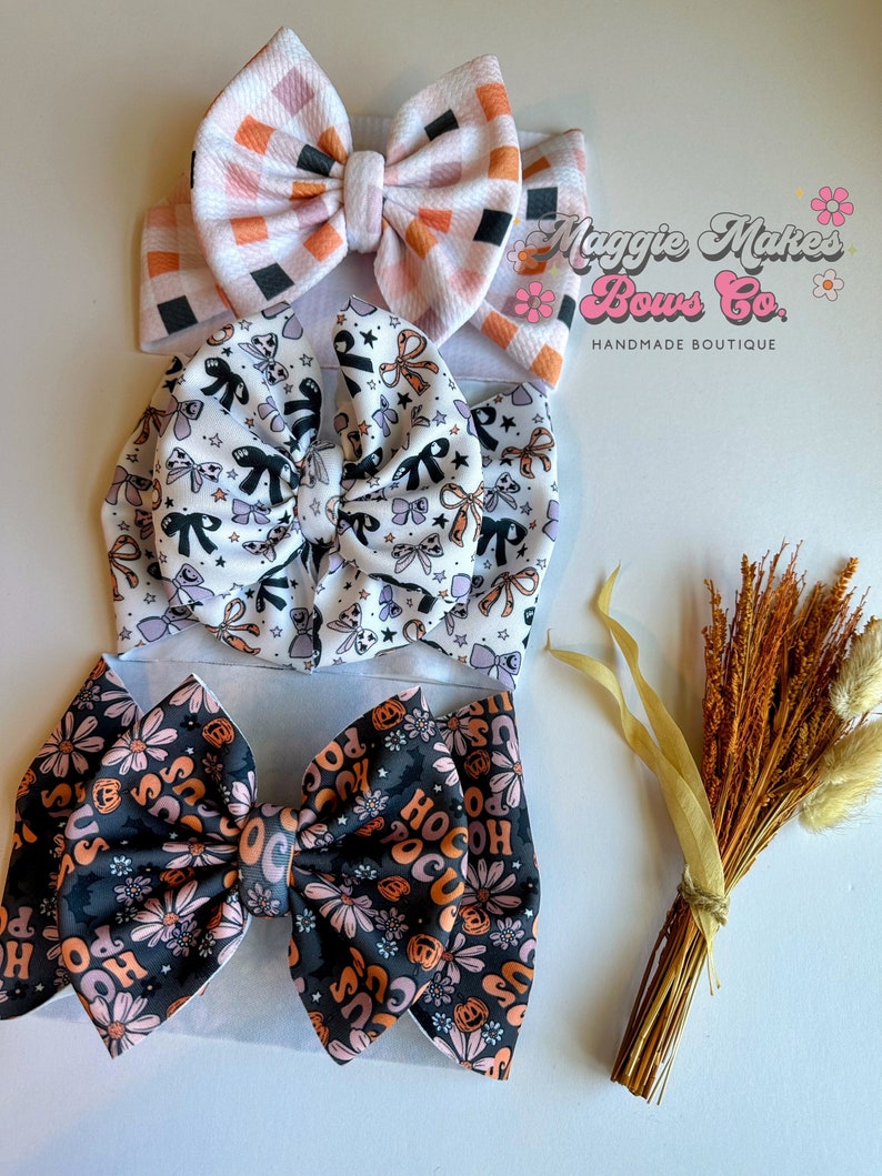 May include: Three fabric headbands with bow accents. The top headband has a pink, orange, and black checkered pattern. The middle headband is white with black and lavender bows. The bottom headband is black with orange flowers and the text "Hocus Pocus".
