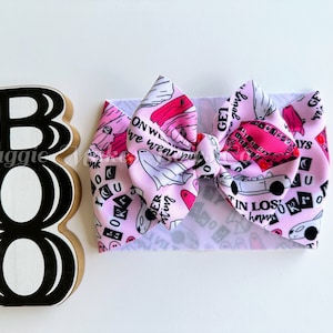 May include: Pink and white fabric hair bow with a Halloween theme. The bow features a ghost, a car, and the text "Get going", "On we wear", "Boo", "Mer rating", "Get in Los", "Always", "You", "Ora", "R5".