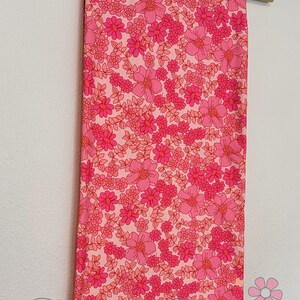 May include: Pink fabric with a floral print featuring pink and red flowers on a white background. The fabric is hanging on a wooden hanger.