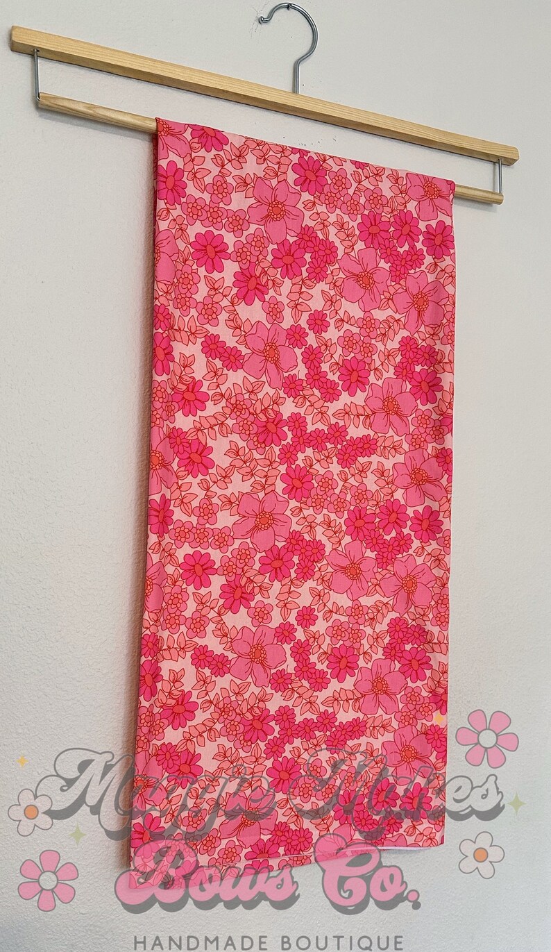 May include: Pink fabric with a floral print featuring pink and red flowers on a white background. The fabric is hanging on a wooden hanger.