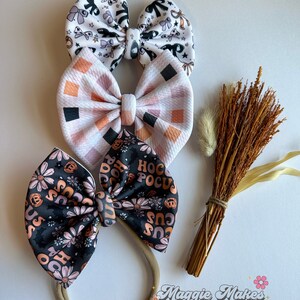 May include: Three handmade fabric hair bows in a variety of patterns. The top bow is white with black and purple Halloween-themed illustrations. The middle bow has a pink and orange checkered pattern. The bottom bow is black with orange flowers and the text "Hocus Pocus".