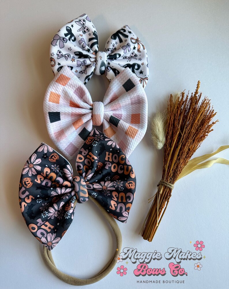 May include: Three handmade fabric hair bows in a variety of patterns. The top bow is white with black and purple Halloween-themed illustrations. The middle bow has a pink and orange checkered pattern. The bottom bow is black with orange flowers and the text "Hocus Pocus".