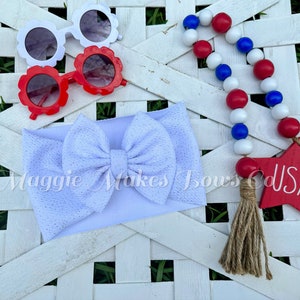 May include: White headband with a white bow, red and white flower-shaped sunglasses, and a red, white, and blue beaded garland with a wooden star that says "USA".